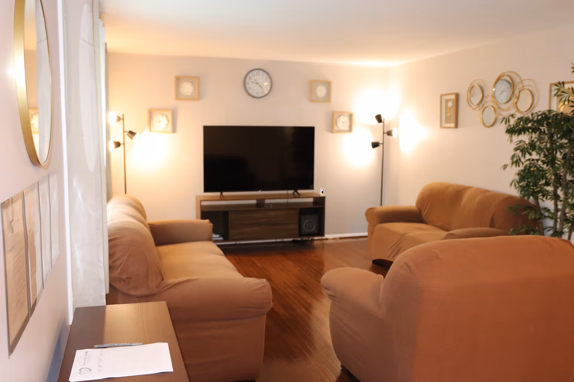 Living room with brown upholstered sofas facing a flat-screen TV on a media console, with floor lamps, wall clocks and decorative wall art.