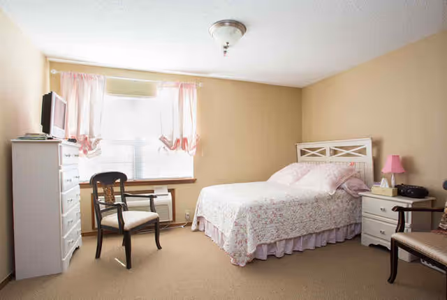 A cozy bedroom with a bed covered in floral bedding, two chairs, a white dresser with a TV on top, a nightstand with a pink lamp, and a window with pink curtains letting in natural light.