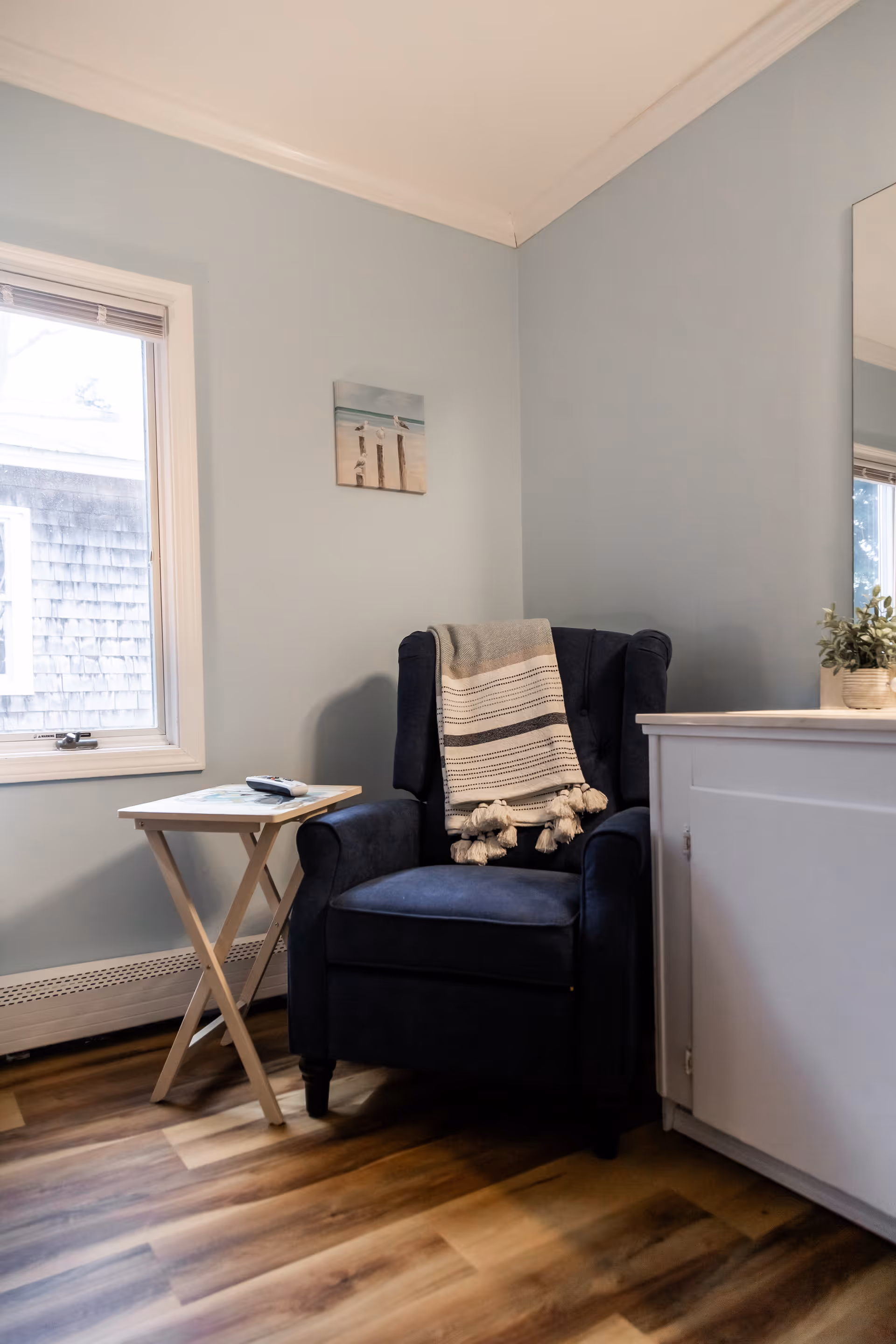 Navy armchair with a striped throw next to a small folding side table in a light-blue corner room with a window and cabinet.