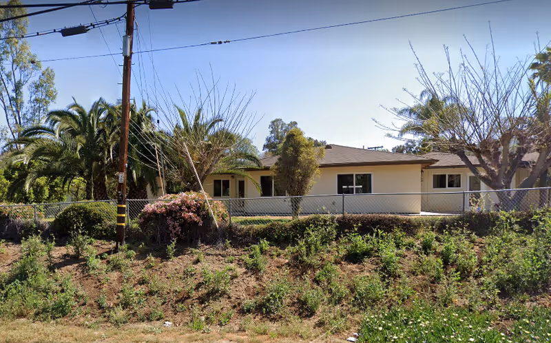 Single-story building with a beige exterior surrounded by trees and bushes, viewed from across a grassy area with a chain-link fence in front. There are palm trees and other greenery around the building under a clear blue sky.