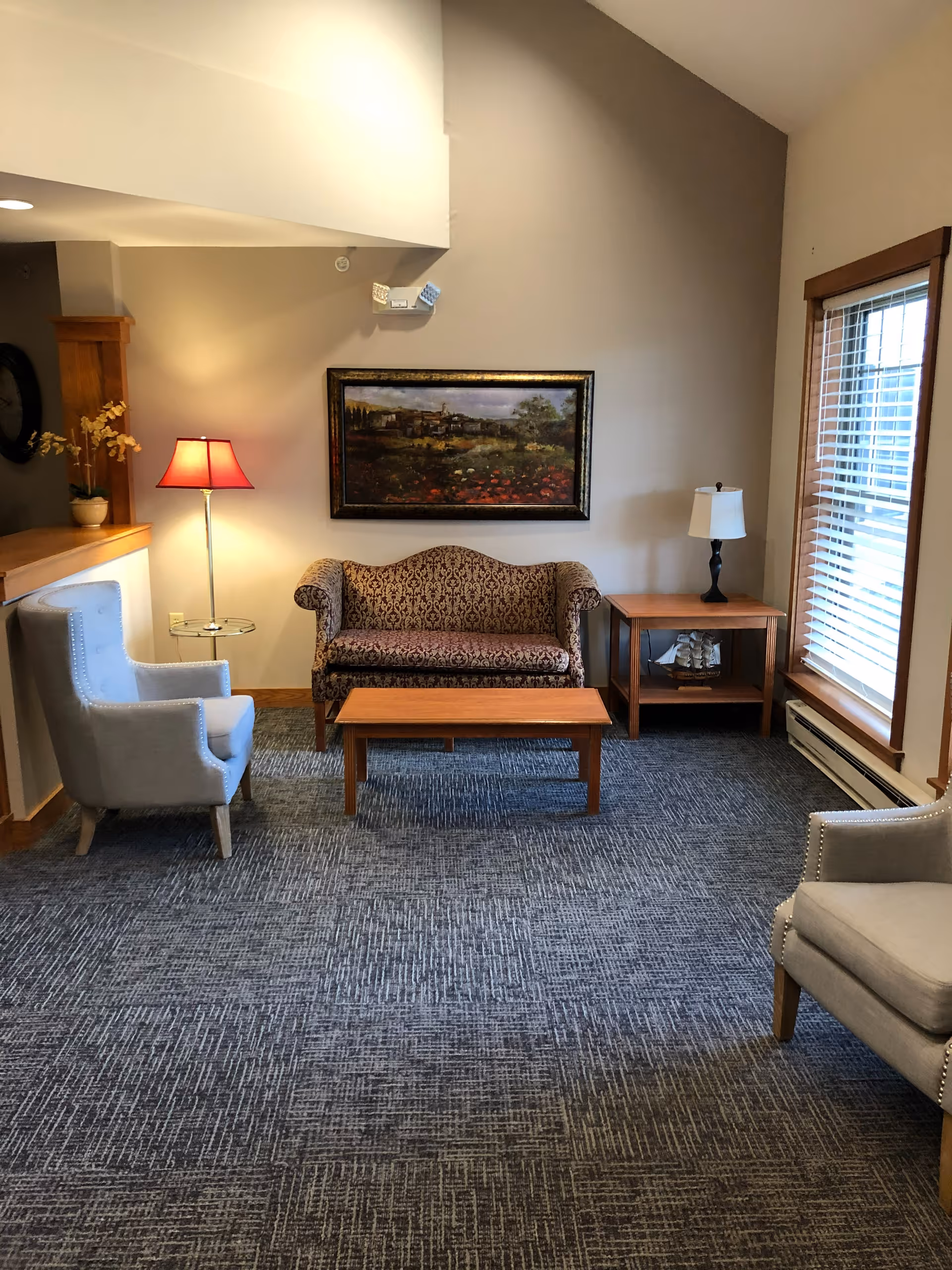 Sitting room with a patterned loveseat, two armchairs, a coffee table, side tables with lamps, a framed landscape painting, and a window with blinds.