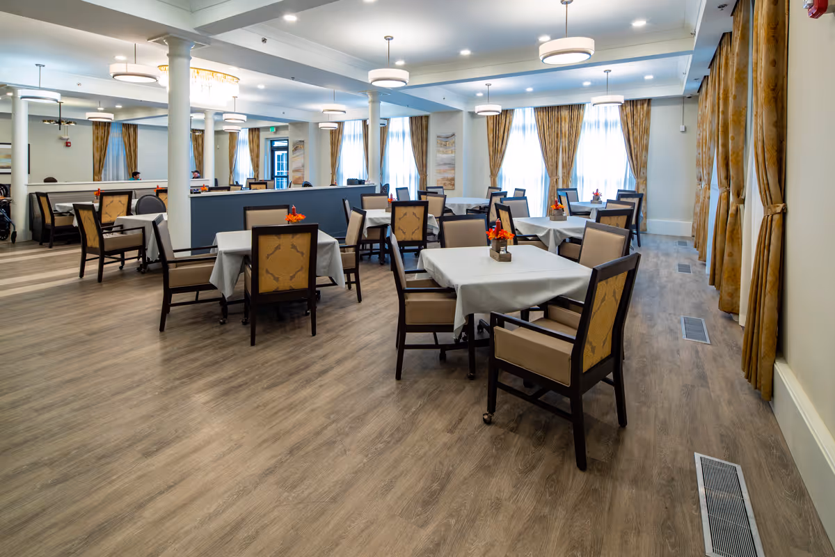 A spacious dining room with multiple tables covered with white tablecloths and surrounded by cushioned chairs. The room has large windows with gold curtains, wooden flooring, white pillars, and modern ceiling lights. Small floral centerpieces decorate each table.