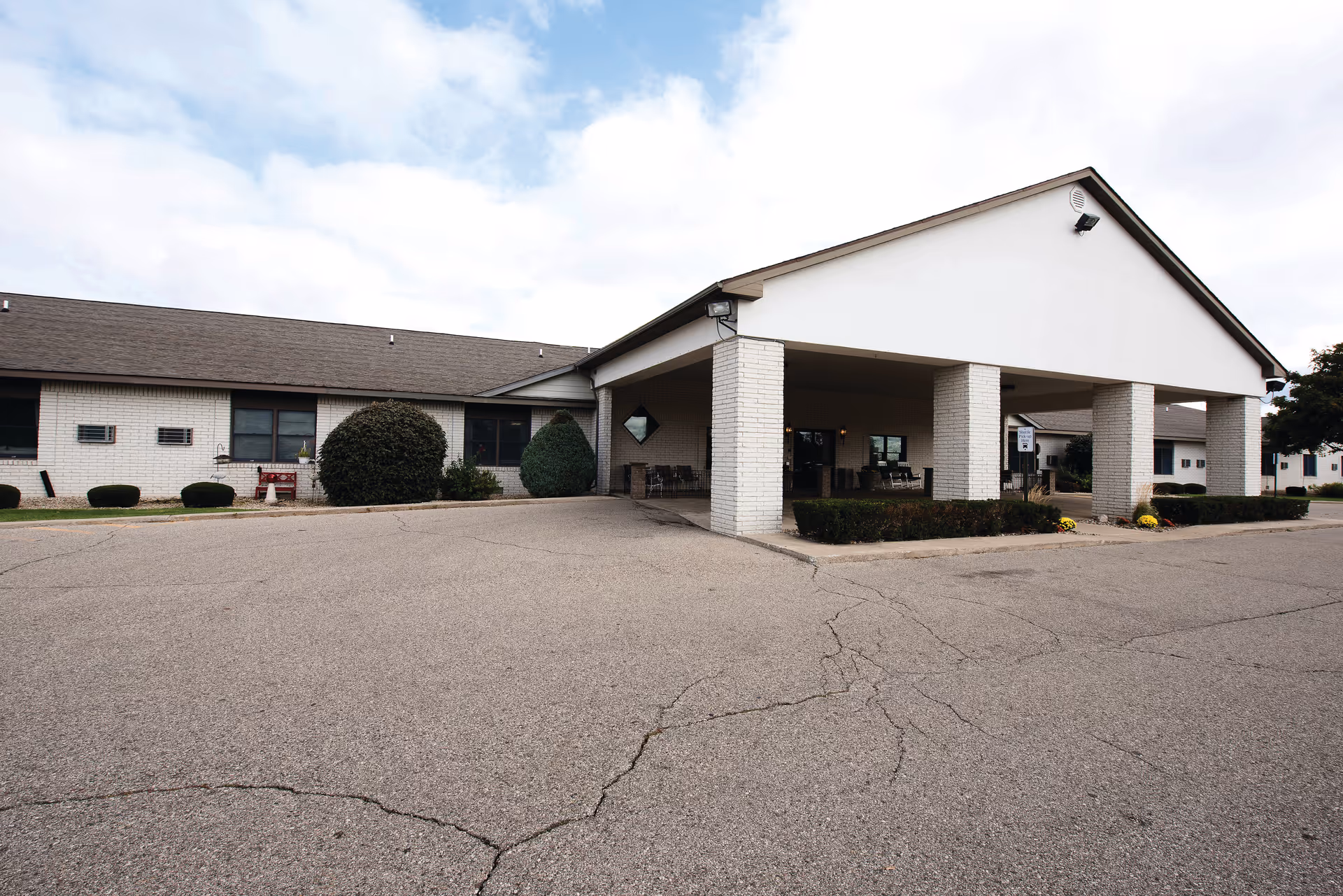 Exterior view of Wellspring Lutheran Services Senior Living Campus - Independent Living building with a covered entrance supported by white brick pillars, surrounded by trimmed bushes and a paved driveway under a partly cloudy sky.