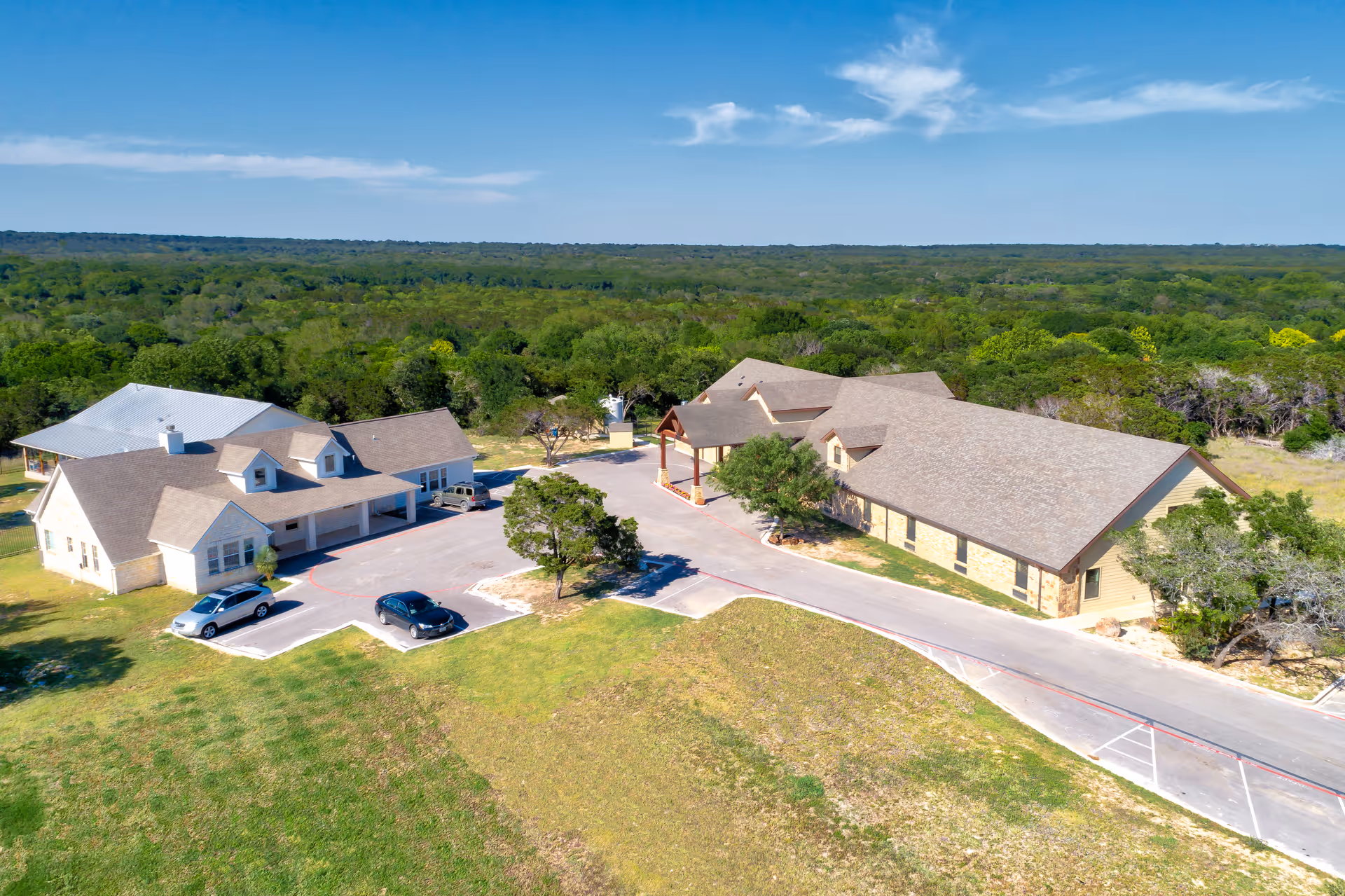 Aerial view of Riva Ridge Assisted Living & Memory Care Center showing multiple connected buildings with beige walls and brown roofs surrounded by green trees and grass under a blue sky.