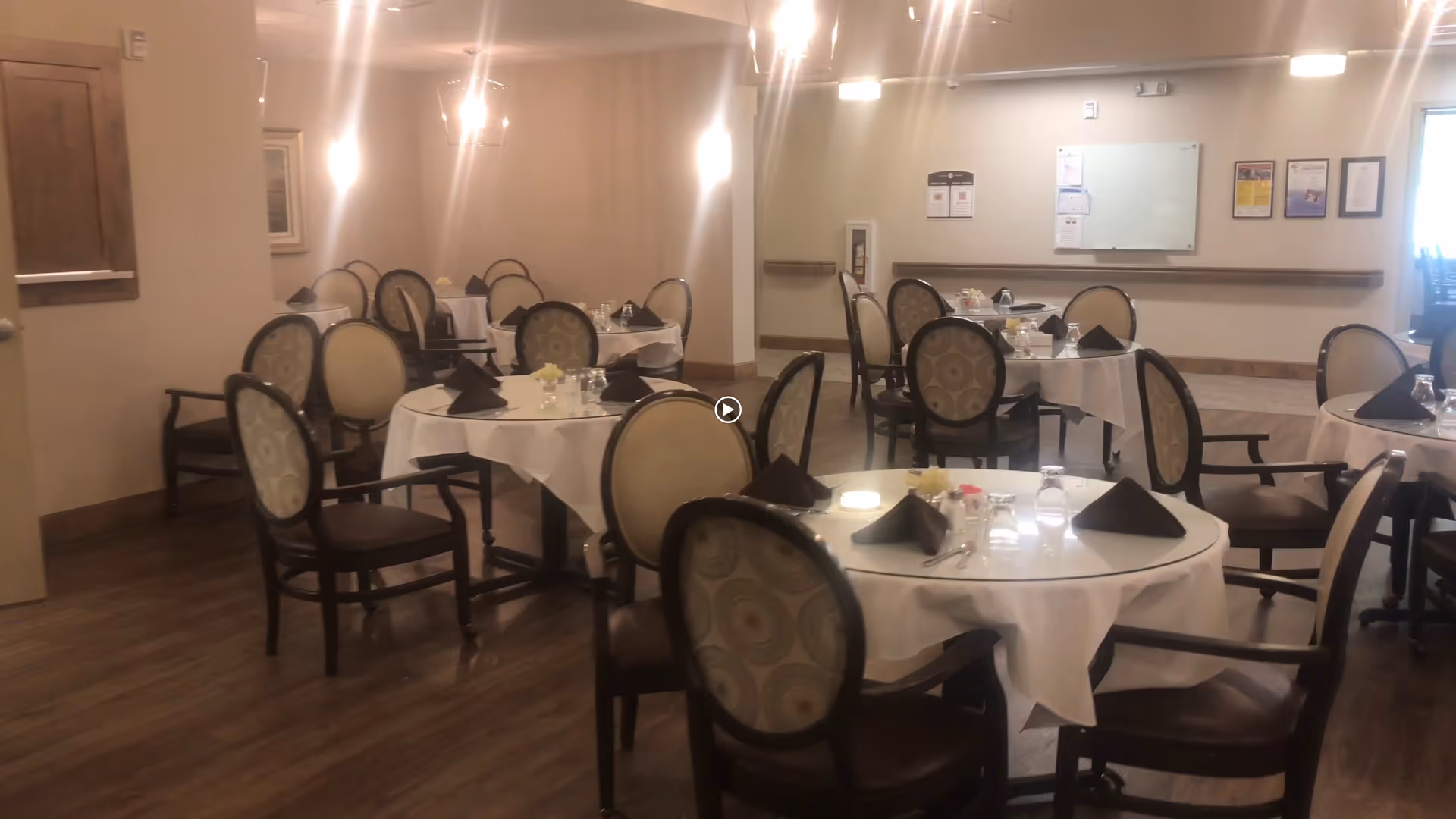 Dining room with round tables covered in white tablecloths, each set with black folded napkins, glasses, and small flower arrangements. The room has wooden flooring, beige walls, and several upholstered chairs around the tables. There are lights hanging from the ceiling and a whiteboard on the far wall.