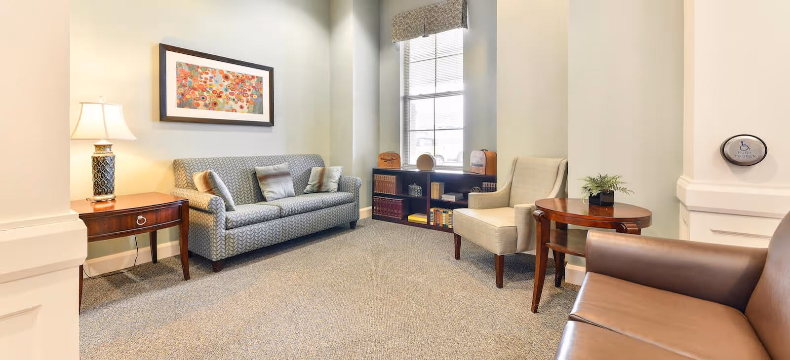A cozy sitting area in a senior living facility with a patterned sofa, an armchair, a brown leather chair, a wooden side table with a lamp, a round wooden table with a small plant, and a bookshelf under a window. The walls are light-colored and there is a framed colorful artwork above the sofa.