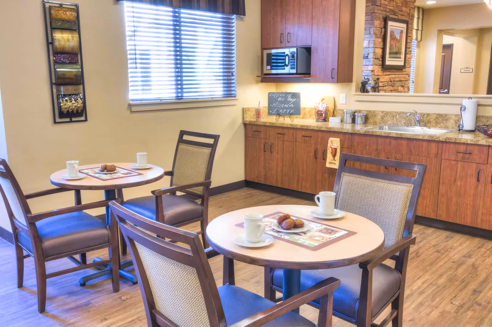 A cozy dining area with two round tables, each set with two chairs. On the tables are cups and plates with muffins. The room has wooden flooring, a window with blinds, and a kitchenette with wooden cabinets, a microwave, a sink, and a small chalkboard sign that reads 'Coffee of the Day: Mocha Latte'.