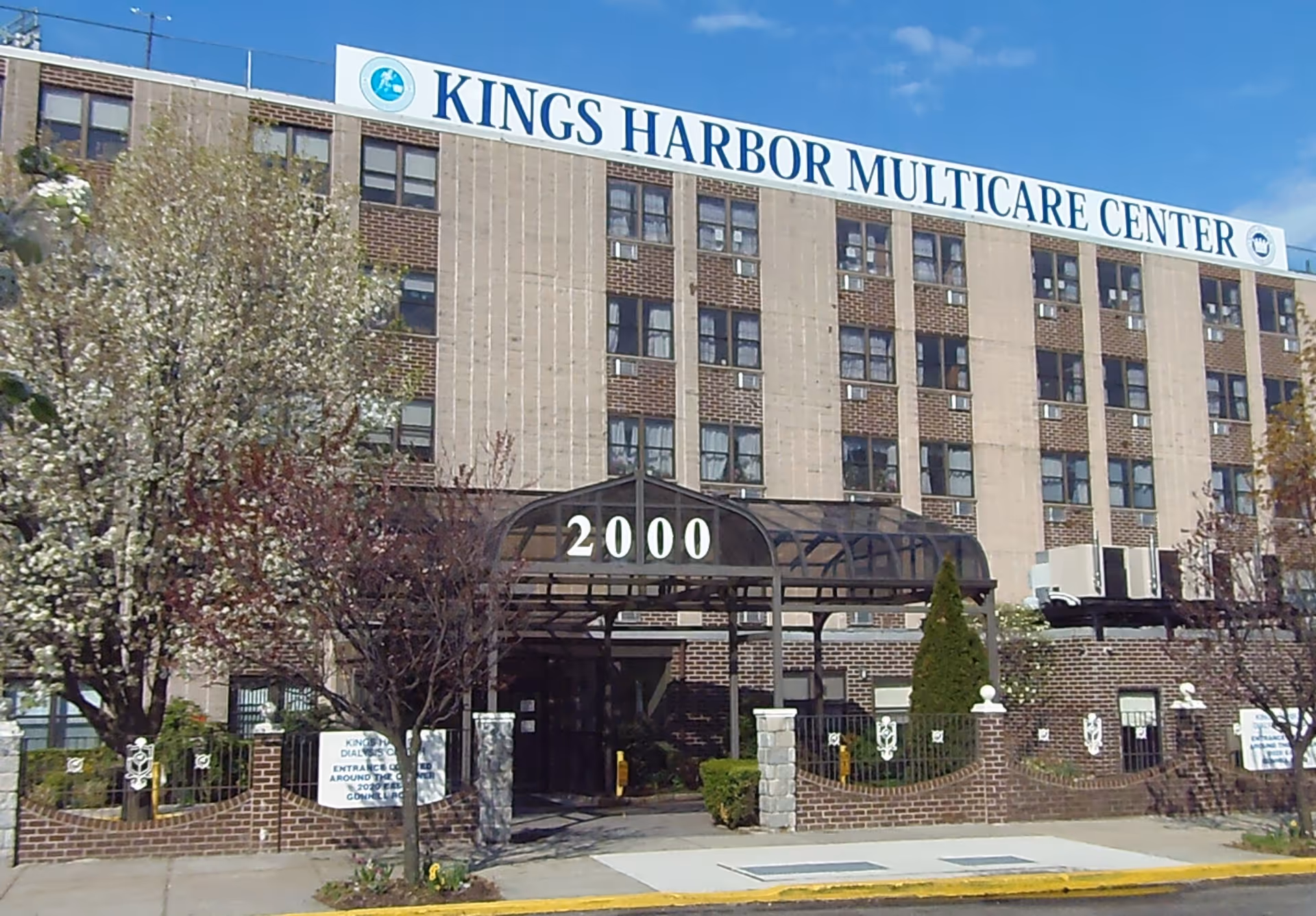 Exterior front view of Kings Harbor Multicare Center, a multi-story brick building with many windows. The entrance is covered with a black metal and glass canopy displaying the number 2000. Trees with blooming flowers and a brick fence are visible in front of the building under a clear blue sky.