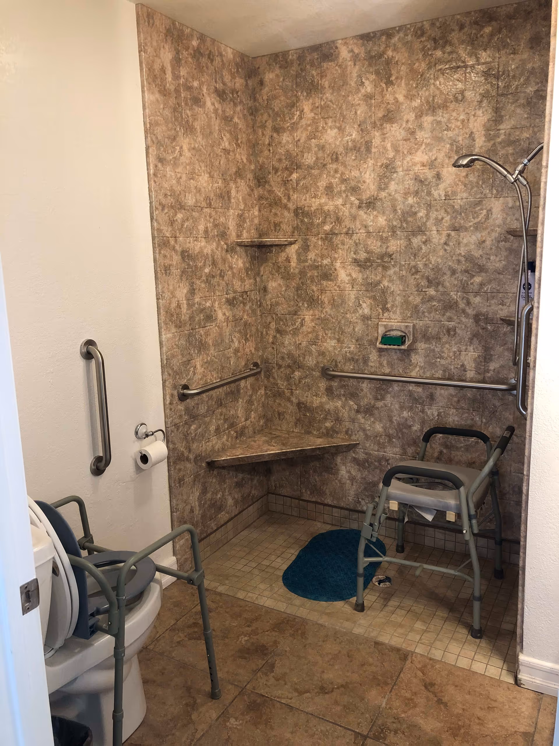 Accessible bathroom with a walk-in shower featuring a built-in corner seat, grab bars on the walls, a handheld showerhead, and a shower chair. There is also a toilet with an elevated seat and armrests, a grab bar on the adjacent wall, and a toilet paper holder.