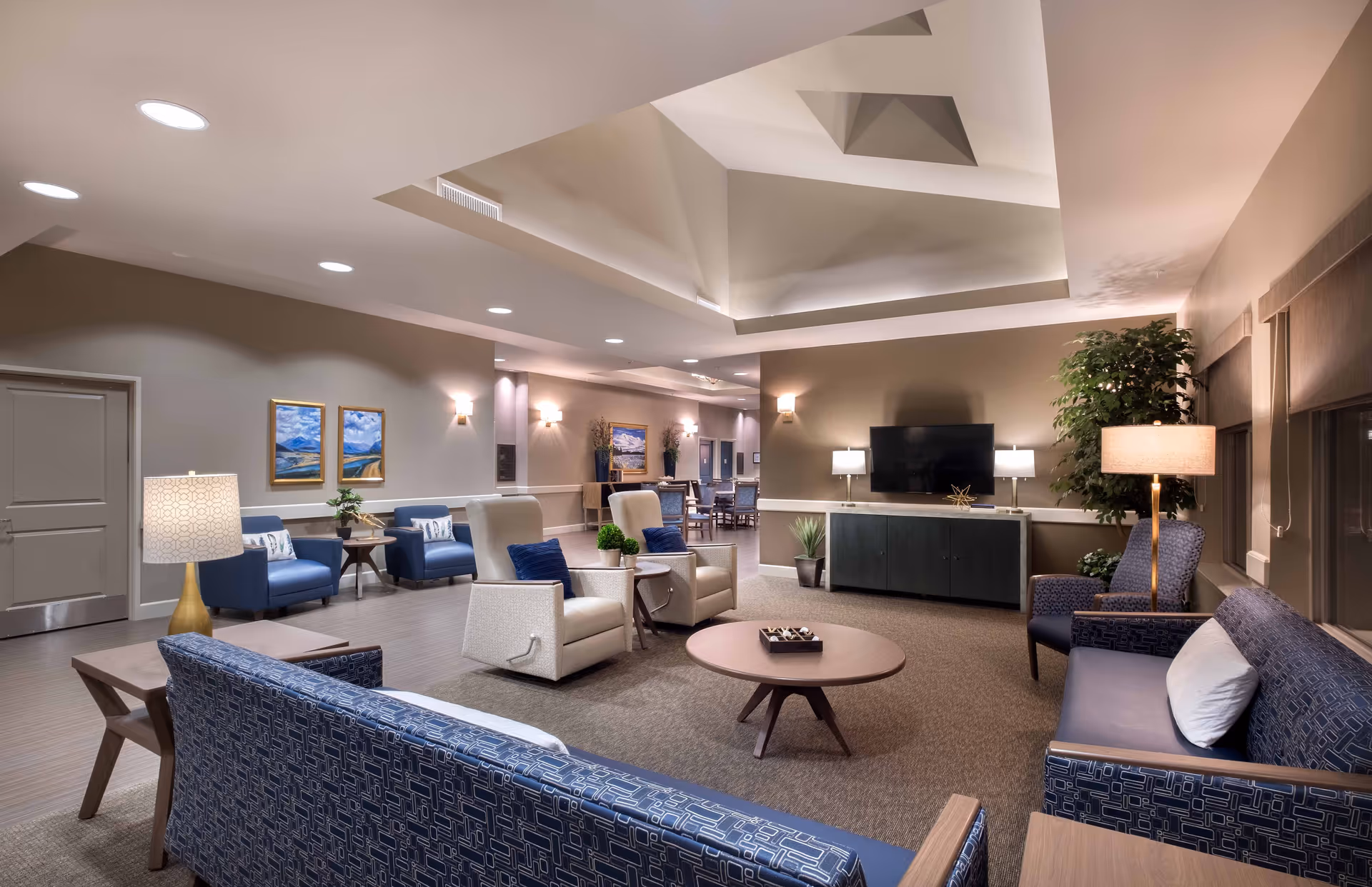 A spacious and well-lit living room area in a senior living facility with modern furniture including blue patterned sofas, beige armchairs with blue cushions, a round wooden coffee table, and side tables with lamps. The room features a high ceiling with recessed lighting, a flat-screen TV mounted on a wall above a dark wooden cabinet, framed landscape paintings on the walls, and a large potted plant near the window.