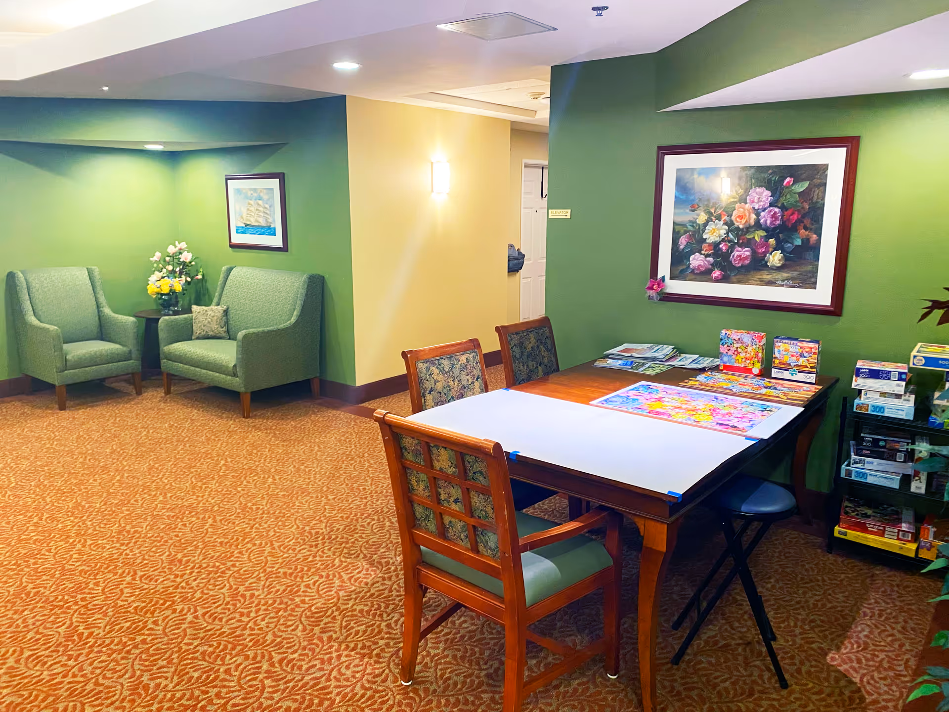 A cozy senior living common area with green walls and patterned carpet. There are two green armchairs and a loveseat with a small round table holding flower arrangements. A wooden table with four chairs is set up with a partially completed colorful jigsaw puzzle and several puzzle boxes. A floral painting hangs on the wall above the table, and a small sign indicating the elevator is visible on the wall.