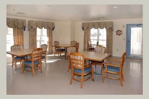 A well-lit dining room with several wooden tables and chairs arranged neatly. The room has large windows with patterned curtains allowing natural light to enter. A blue door with a window is visible on the right side, and a decorative ship wheel is mounted on the wall nearby.