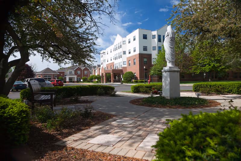 Outdoor view of a senior living facility with a paved walkway, a statue on a pedestal, benches, trees, bushes, and a multi-story building in the background under a partly cloudy sky.