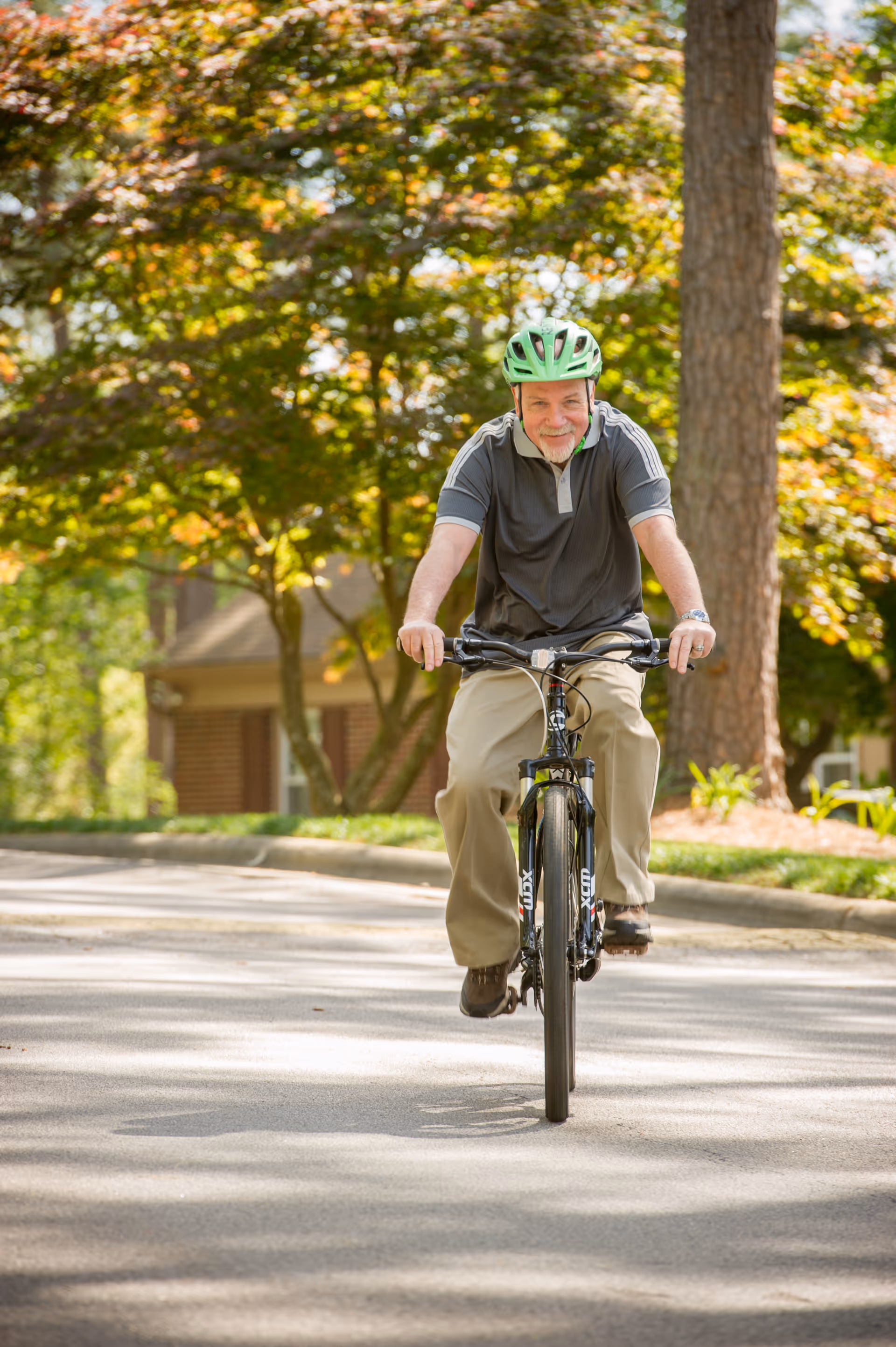 An elderly man wearing a green helmet and casual clothes rides a bicycle on a paved path surrounded by trees and greenery in a residential area.