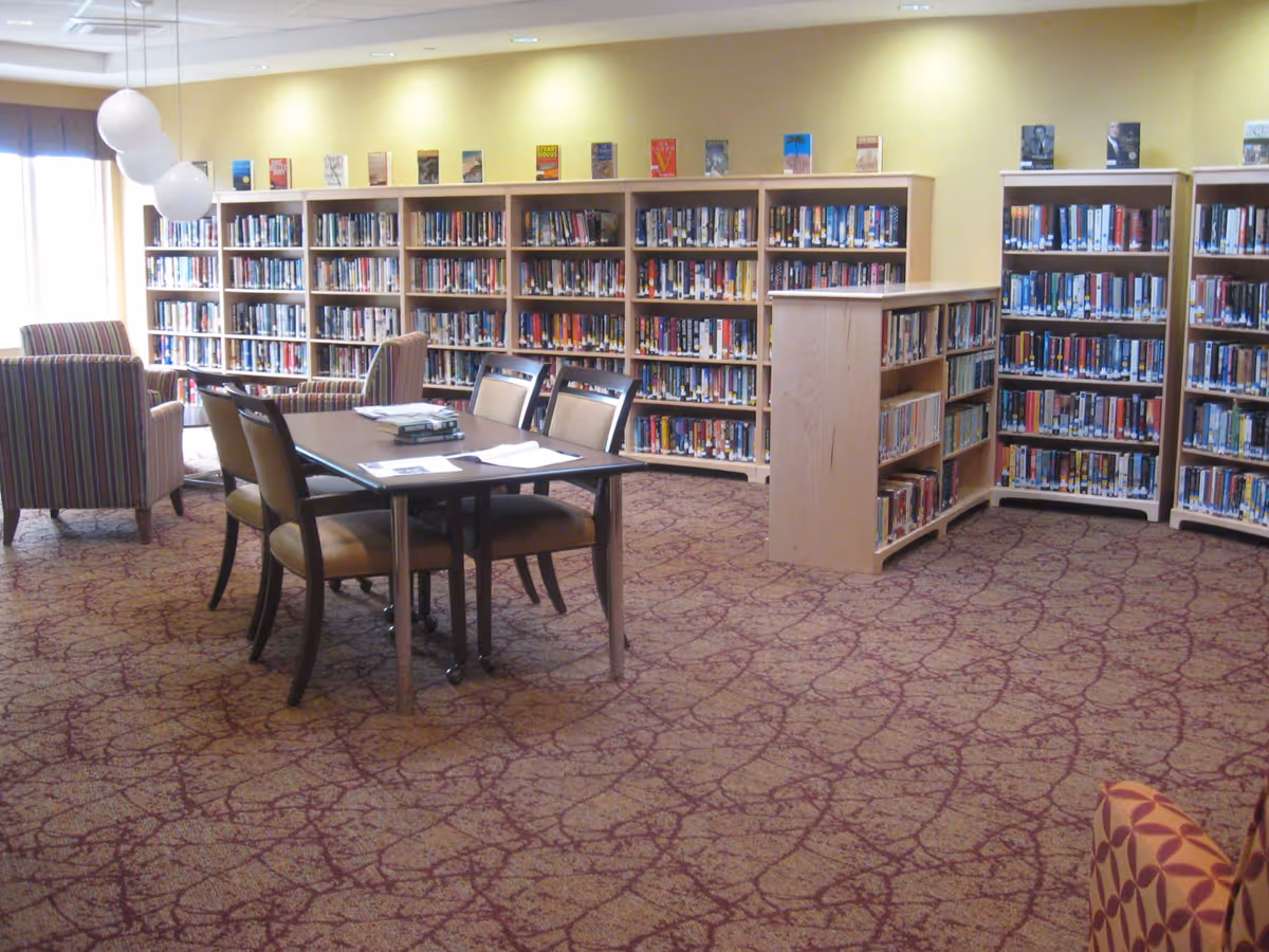 A well-lit library room with multiple bookshelves filled with books along the walls and a central bookshelf. There is a table with four chairs in the middle of the room, and two striped armchairs near the window. The carpet has a patterned design, and the walls are painted a soft yellow.