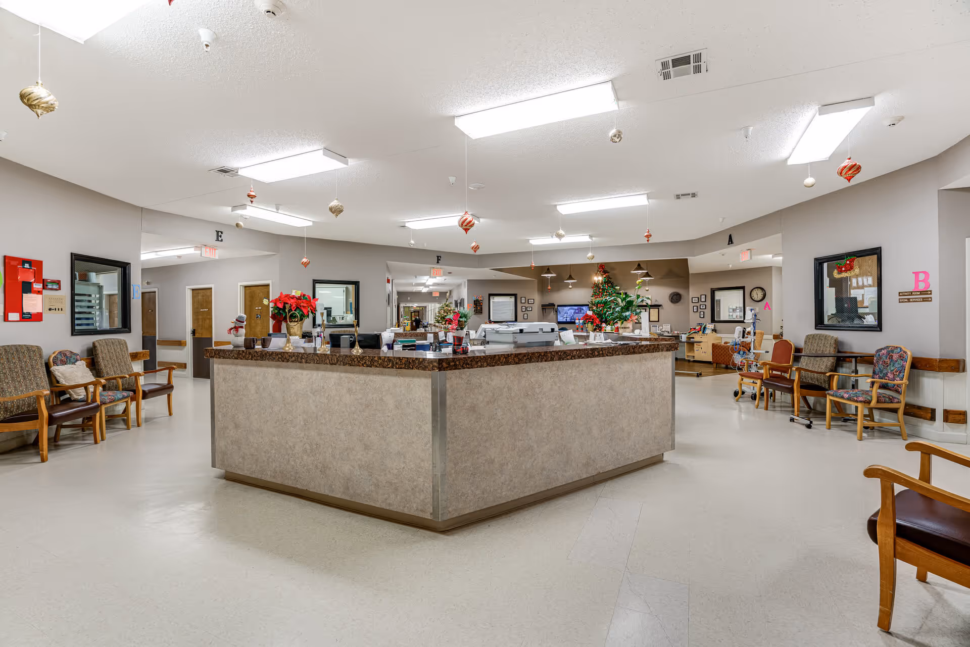 A spacious senior living facility reception area with a large central desk decorated with poinsettias and holiday ornaments hanging from the ceiling. Surrounding the desk are several chairs with cushions, and the walls have mirrors and signs indicating different sections labeled A, B, E, and F. The area is well-lit with ceiling lights and has a clean, welcoming atmosphere.