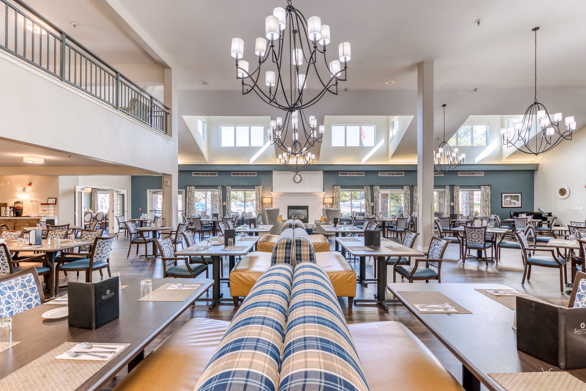 Spacious dining room in a senior living facility with multiple tables and chairs arranged neatly. The room features high ceilings with large chandeliers, windows allowing natural light, and a fireplace at the far end. Tables are set with placemats, utensils, glasses, and menus. The seating includes cushioned benches with blue plaid upholstery and individual chairs with patterned fabric.