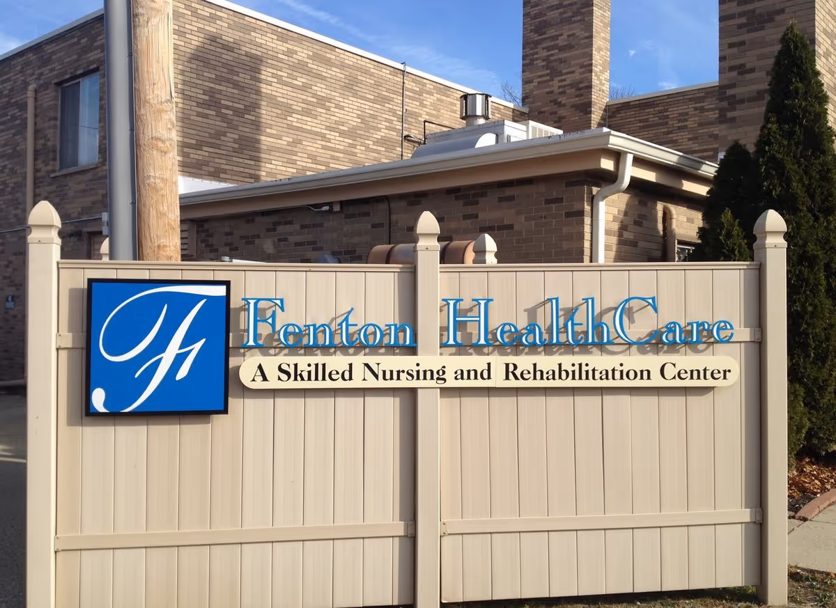 Sign for Fenton HealthCare, a Skilled Nursing and Rehabilitation Center, mounted on a beige fence with a brick building and trees in the background.
