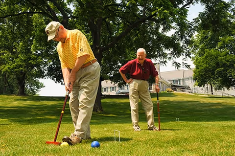 Two elderly men playing croquet on a grassy lawn with trees and buildings in the background on a sunny day.