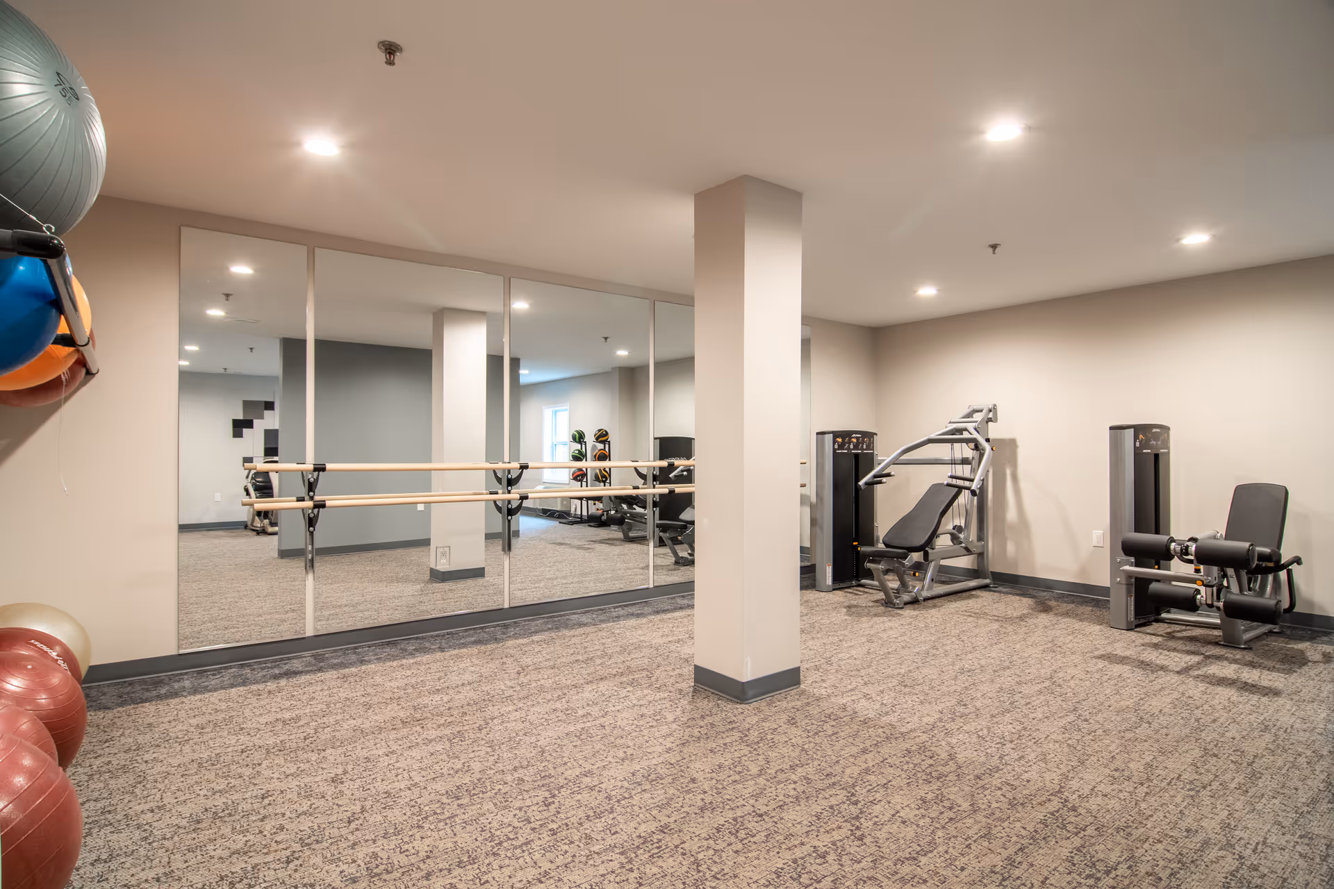 A fitness room with exercise equipment including a leg press machine and a leg extension machine. There are large mirrors on one wall with ballet barre rails attached. Exercise balls are stored on a rack mounted on the wall. The room has carpeted flooring and neutral-colored walls with recessed lighting.