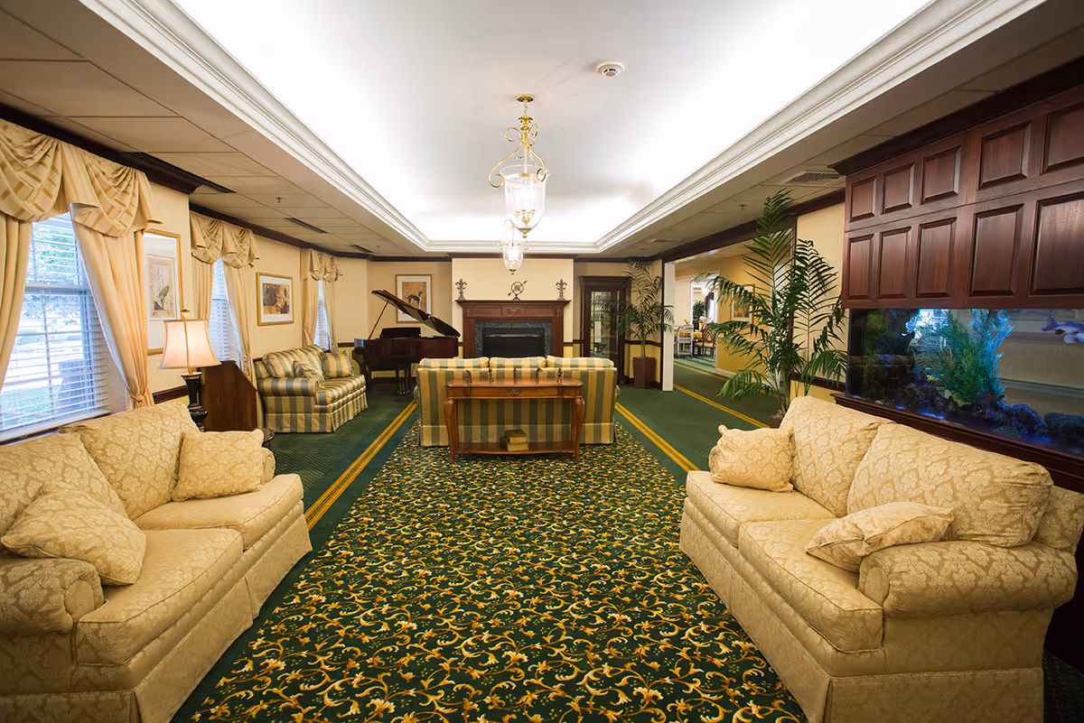 Spacious senior living lounge with sofas, patterned green carpet, a fireplace, grand piano, and an aquarium.
