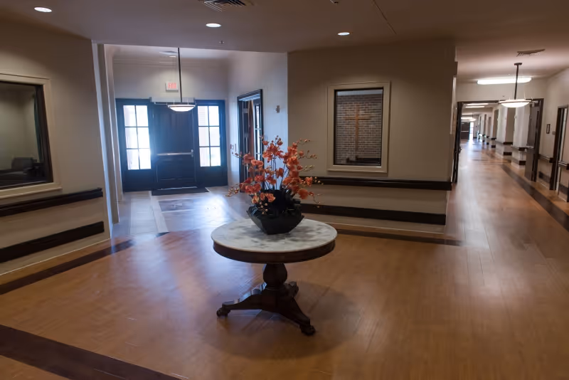 Interior view of a hallway in a healthcare facility with wooden flooring, a round table with a flower arrangement in the center, and double doors with windows at the end. The hallway extends to the right with ceiling lights and handrails along the walls.