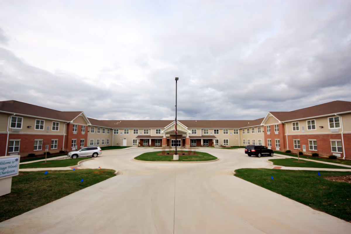 Front exterior view of Heritage Woods of Charleston, a two-story senior living facility with beige and red brick walls, a central entrance with a covered porch, an American flag on a pole in the circular driveway, and two parked cars on either side.