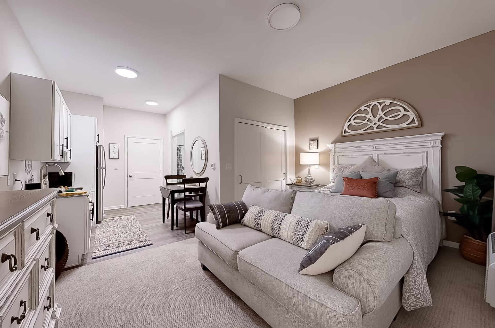 A cozy studio apartment featuring a beige sofa with decorative pillows in the foreground, a white wooden bed with multiple pillows and a decorative wall piece above the headboard, a bedside table with a lamp, and a small dining table with two chairs near a compact kitchen area with white cabinets and stainless steel appliances.