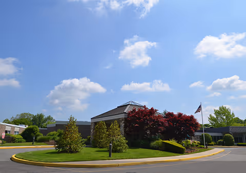Exterior view of Carillon Nursing and Rehabilitation Center showing a single-story building with a landscaped circular driveway, green trees, bushes, and an American flag on a flagpole under a partly cloudy blue sky.