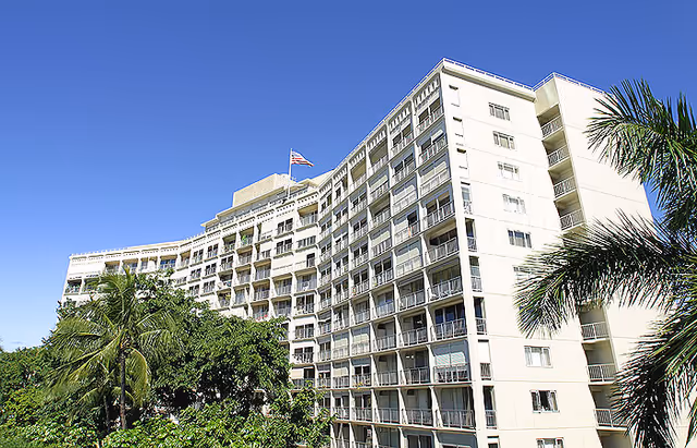Exterior view of a multi-story retirement residence building with balconies, surrounded by palm trees and greenery under a clear blue sky.
