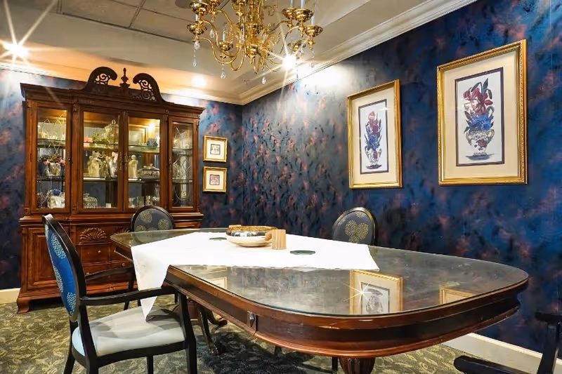 A formal dining room with a dark wooden table covered partially by a white table runner. Four upholstered chairs surround the table. A wooden china cabinet filled with glassware and decorative items stands against a textured dark blue wall. Two framed floral paintings hang on the wall, and a gold chandelier with crystal accents hangs from the ceiling.
