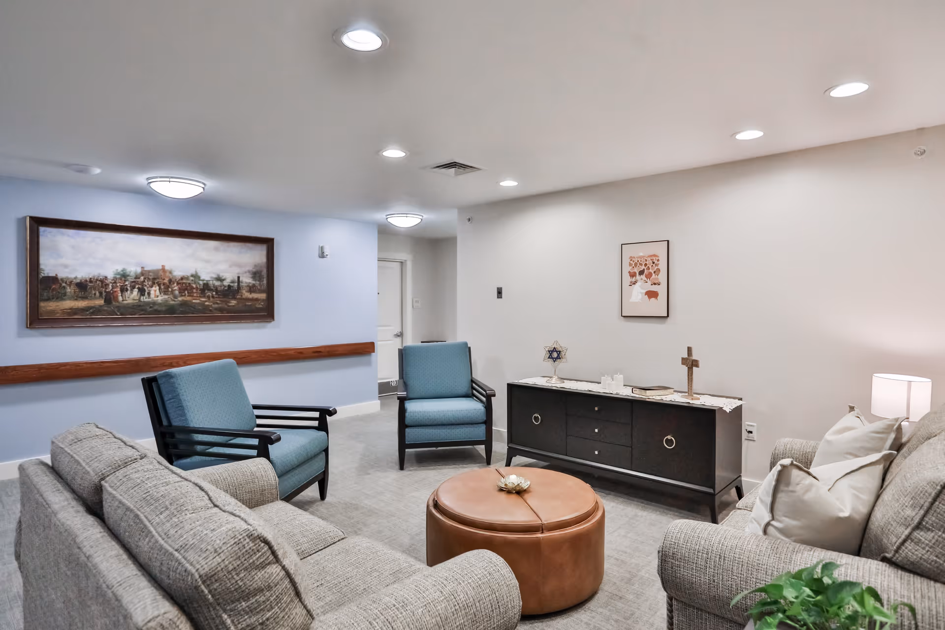 A cozy living room area in a senior living facility featuring two gray upholstered sofas with pillows, two blue armchairs, and a round brown leather ottoman in the center. The walls are painted light blue and white, adorned with framed artwork. A black sideboard with decorative items including a cross and a Star of David sits against one wall. The room is softly lit by ceiling lights and a table lamp.