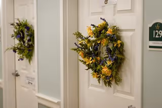 Close-up view of two white doors in a senior living facility hallway, each decorated with a floral wreath featuring yellow and purple flowers. One door has a room number plaque labeled 129 and a nameplate below it.