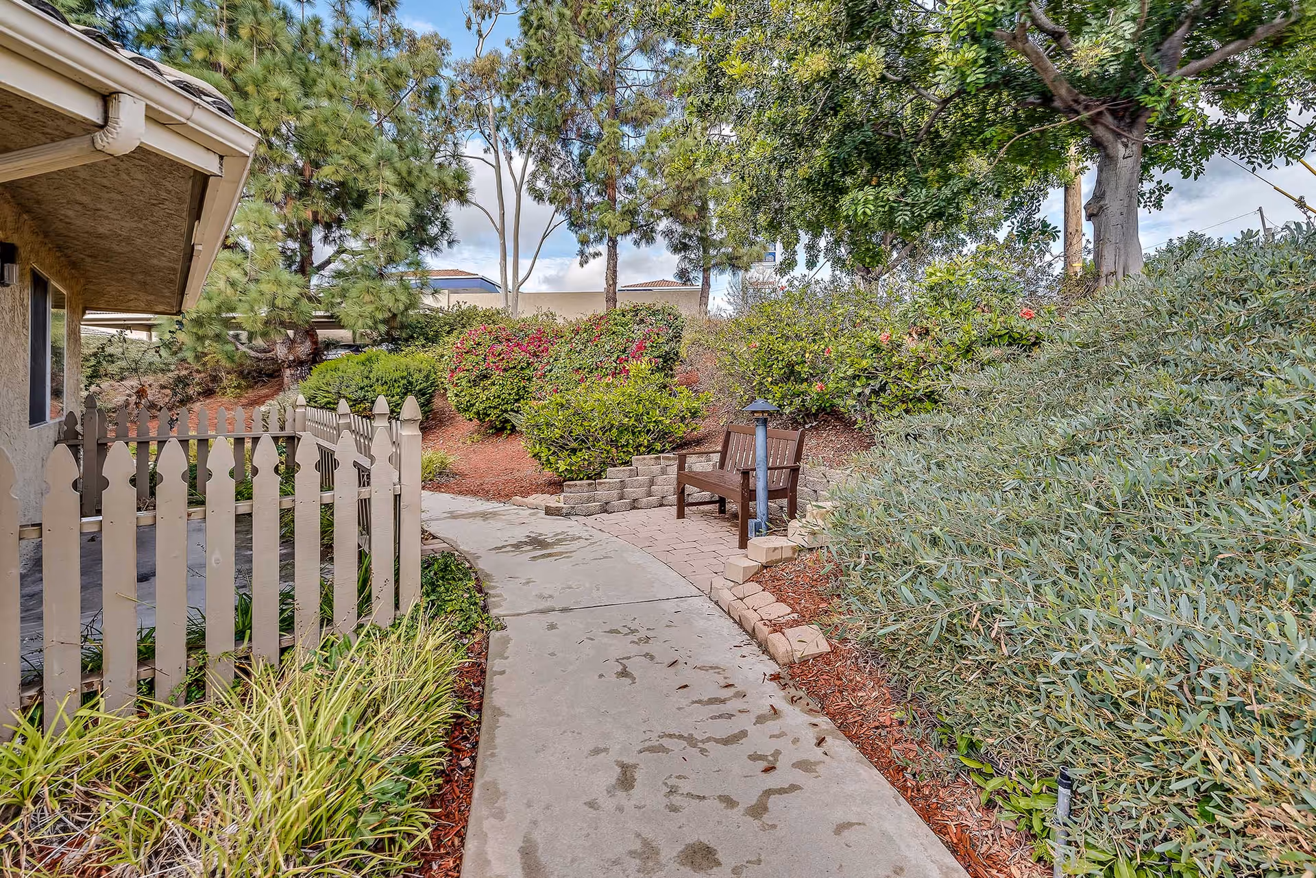 A paved outdoor walkway surrounded by lush greenery and bushes, with a wooden bench and a small lamp post on the right side. A beige picket fence and part of a building are visible on the left side under a partly cloudy sky.