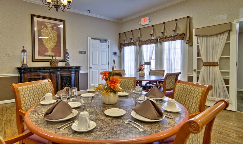 Round dining table set for a meal with folded napkins, dishes, and a floral centerpiece in a warmly decorated dining room.