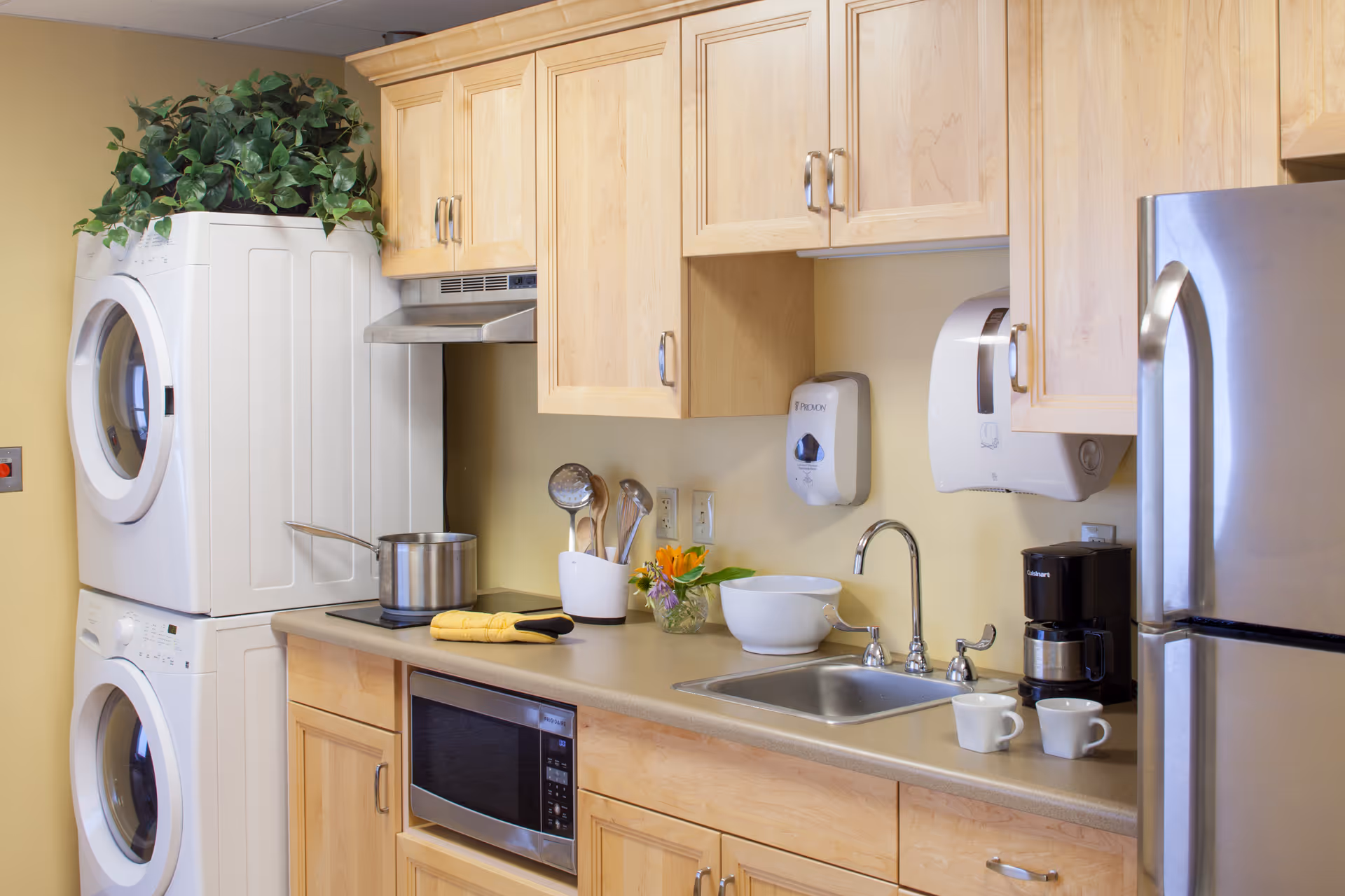 A compact kitchen area with light wood cabinets, a stainless steel refrigerator, microwave, stovetop with a pot, a sink, coffee maker with two white mugs, and a stacked washer and dryer. There is a small plant on top of the dryer and a small flower arrangement on the counter.
