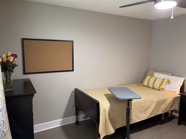A small bedroom with a single bed covered in a yellow quilt and a yellow striped pillow. There is a small adjustable overbed table on wheels positioned over the bed. A black dresser with a vase of colorful flowers is on the left side, and a cork bulletin board is mounted on the wall above it. The walls are painted light gray, and a ceiling fan with a light is visible above.