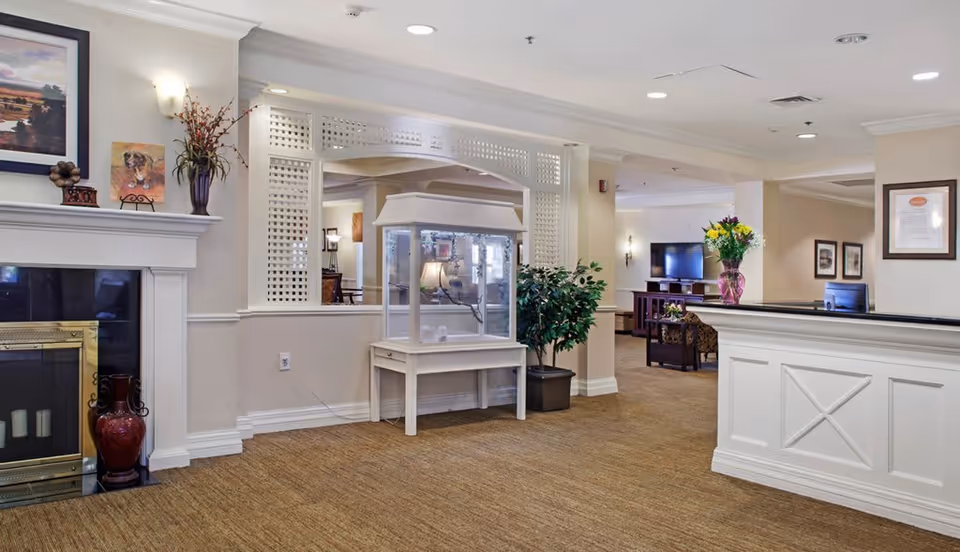 Interior view of a senior living facility lobby area with a white reception desk on the right, a decorative fireplace on the left, a display case in the center, and a seating area with a television in the background. The space is decorated with plants, flowers, framed pictures, and warm lighting.