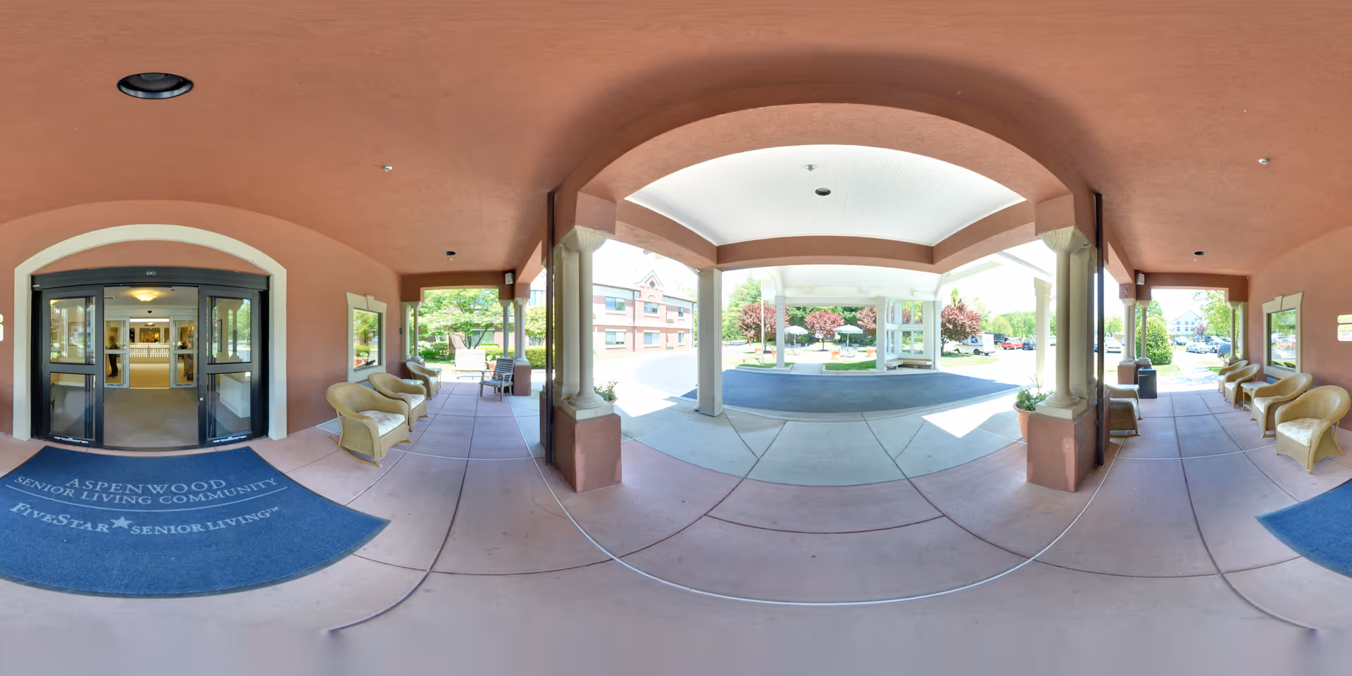 Covered entrance area of Aspenwood Senior Living Community with a blue mat displaying the facility's name and slogan. The area has several wicker chairs arranged along the walls under a pinkish-brown ceiling. The entrance doors lead inside the building, and outside the covered area, there is a driveway and landscaped grounds with trees and shrubs.