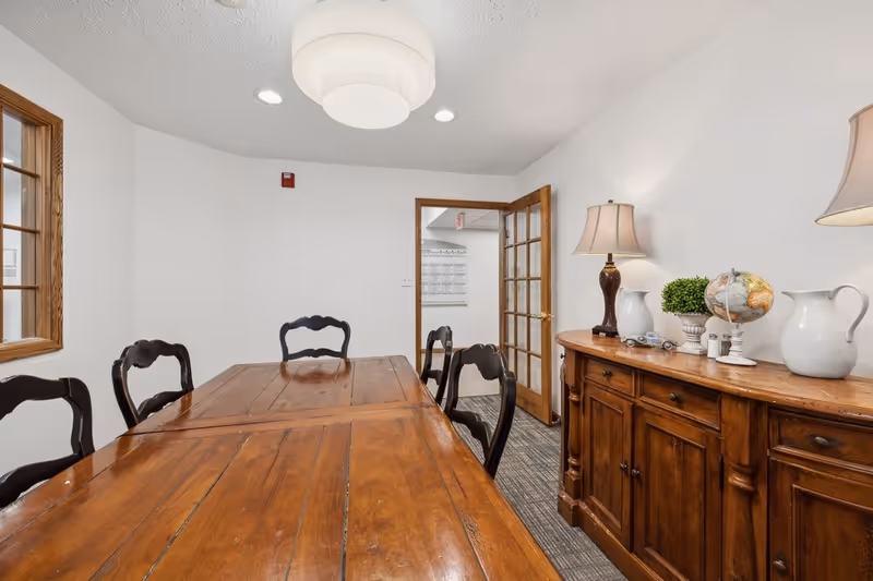 A small meeting or dining room with a long wooden table surrounded by six dark wooden chairs. There is a wooden sideboard on the right side with decorative items including a lamp, a small globe, a plant, and white pitchers. The walls are white, and there is a glass-paneled wooden door leading to another area. A round ceiling light fixture is visible above the table.