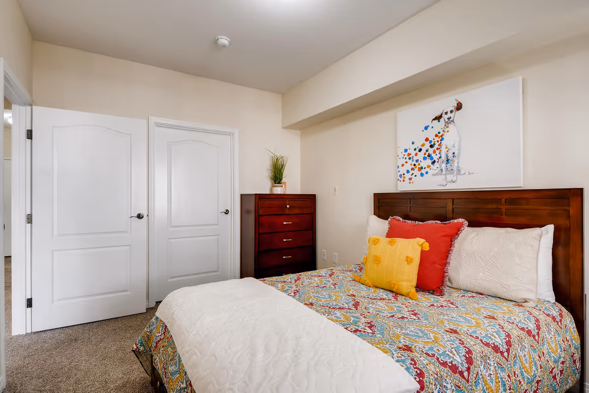Bedroom with a patterned bedspread and pillows, wooden headboard, chest of drawers, two closed white doors, and a dog painting above the bed.