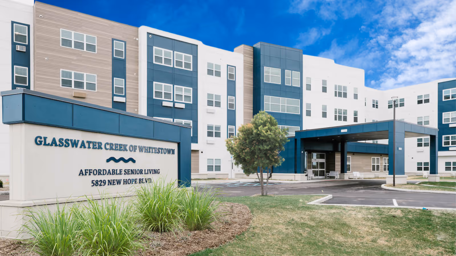 Exterior view of Glasswater Creek of Whitestown, a modern senior living facility with a multi-story building, a covered entrance, and a sign in front that reads 'Glasswater Creek of Whitestown Affordable Senior Living 5829 New Hope Blvd.' The sky is blue with some clouds, and there is landscaping with grass and plants around the sign.