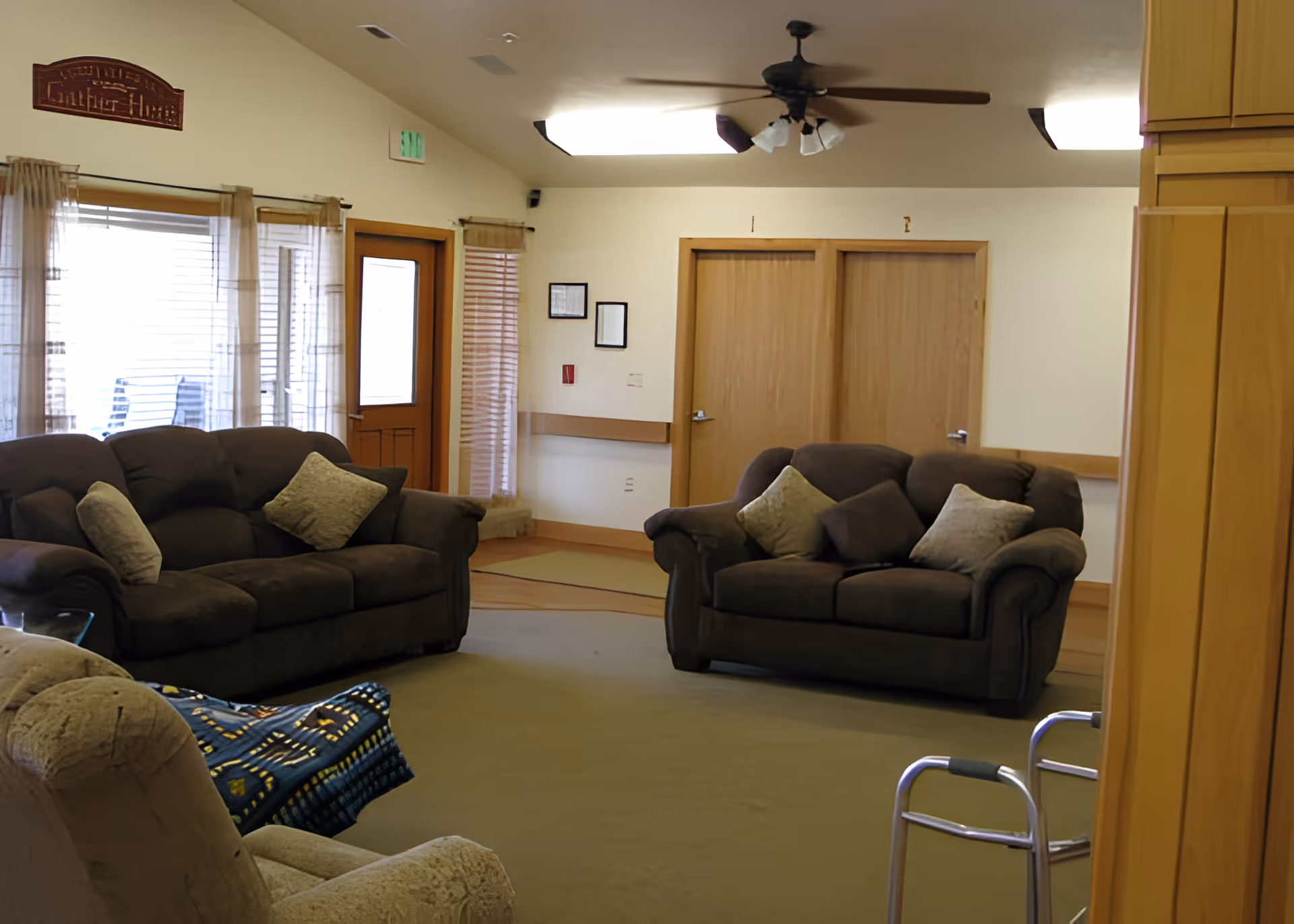 A cozy living room with two dark brown sofas and a beige armchair. The sofas have several cushions. There is a ceiling fan with lights above, wooden doors and windows with blinds and sheer curtains, and a walker is visible in the foreground. The room has a warm, inviting atmosphere with neutral-colored walls and carpet.
