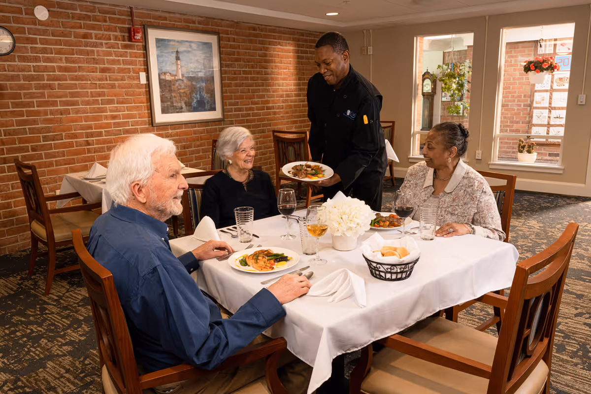 Three elderly people sitting at a dining table in a restaurant-style room with a white tablecloth, enjoying a meal served by a waiter. The table has plates of food, glasses of wine and water, a basket of bread rolls, and a white floral centerpiece. The room has brick walls, framed artwork, and windows letting in natural light.