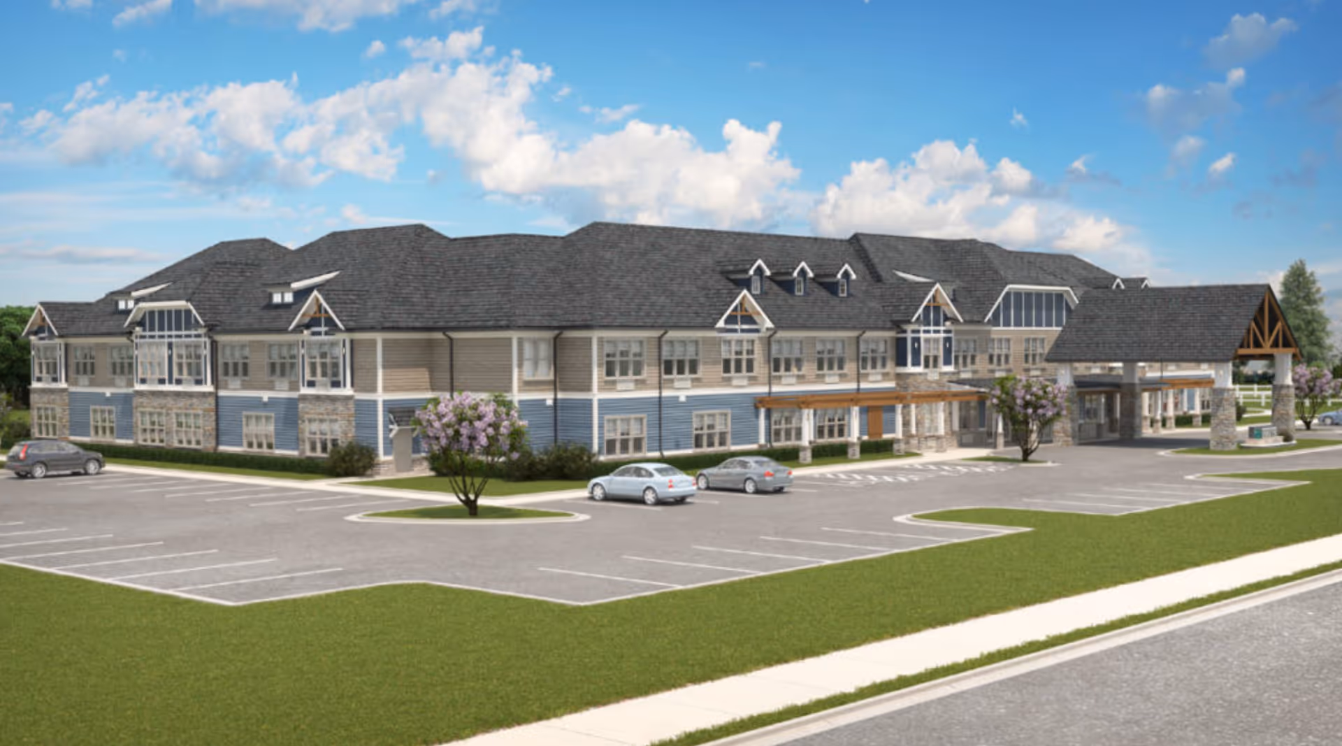Exterior view of a large two-story senior living facility building with a covered entrance, multiple windows, and a spacious parking lot with a few cars parked. The building has a combination of beige and blue siding with stone accents and a dark shingled roof. The sky is blue with scattered clouds.