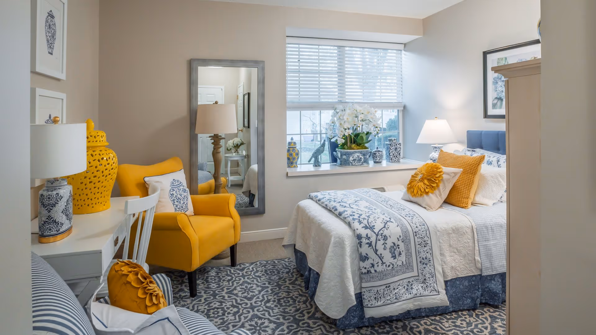 A cozy bedroom with a bed dressed in white and blue floral bedding, accented with yellow and beige pillows. There is a yellow armchair with a white pillow, a white desk with a blue and white lamp and decorative yellow vase, a large mirror on the wall, and a window with white blinds letting in natural light. The room has a patterned blue and white rug and neutral-colored walls.