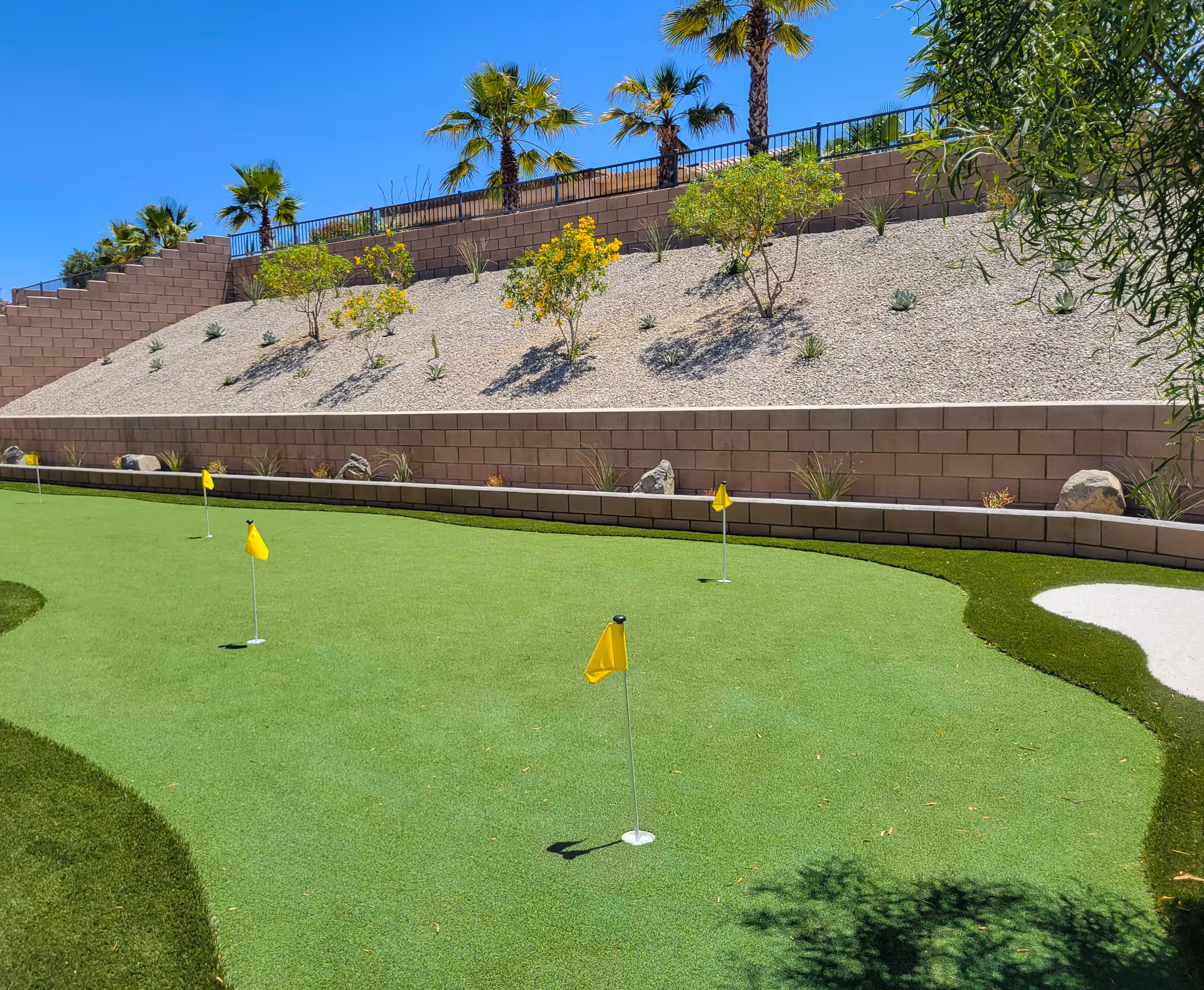 Outdoor putting green with several yellow flags, bordered by a retaining wall, palm trees, and desert landscaping under a clear blue sky.