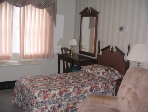 A bedroom with a single bed covered in a floral bedspread, a wooden headboard, a wooden vanity with a mirror and a chair, two table lamps, a beige armchair, and a window with pink curtains.