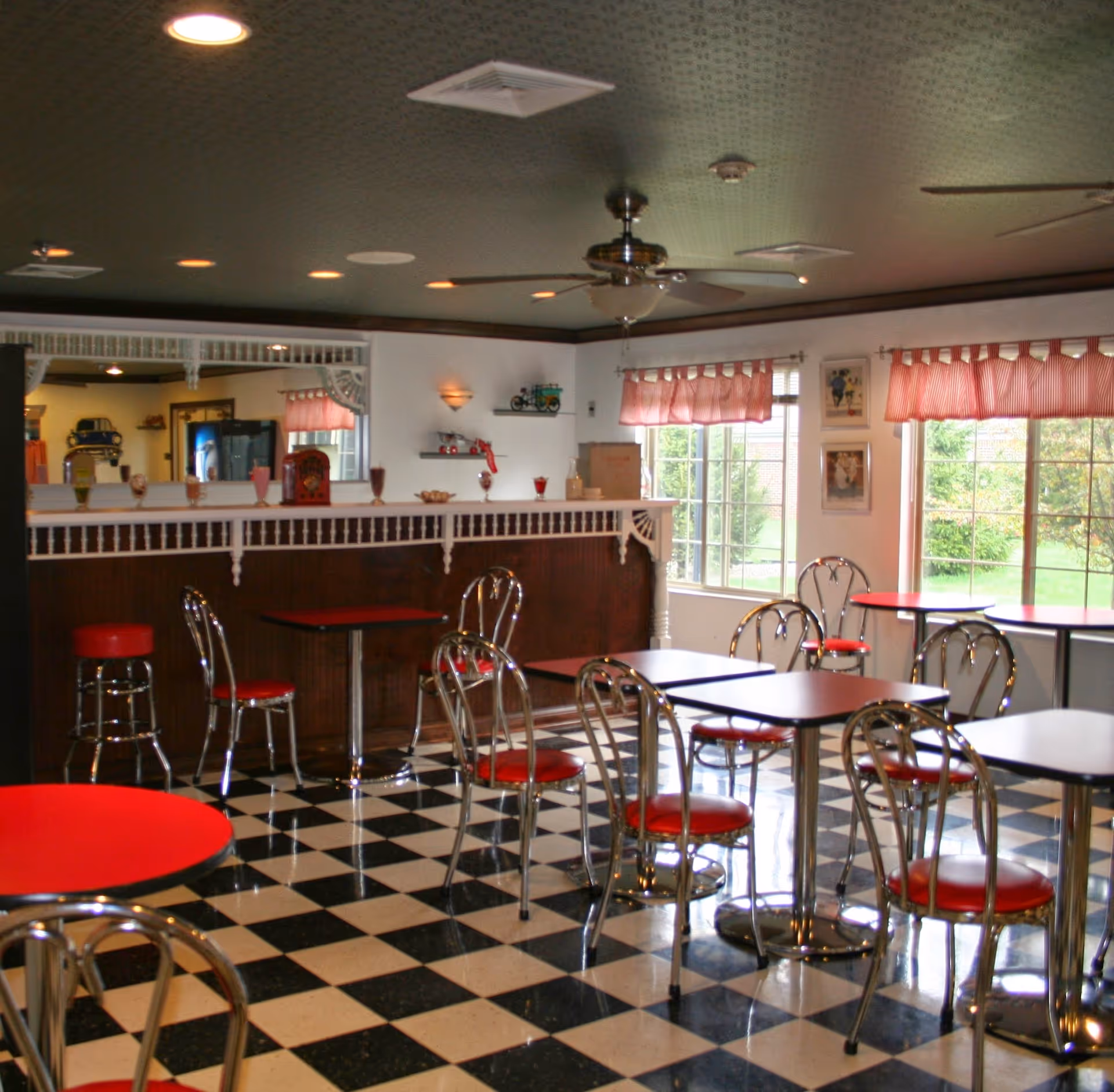 Bright retro-style dining room with black-and-white checkered floor, red-and-chrome tables and chairs, a serving counter and large windows with valances.
