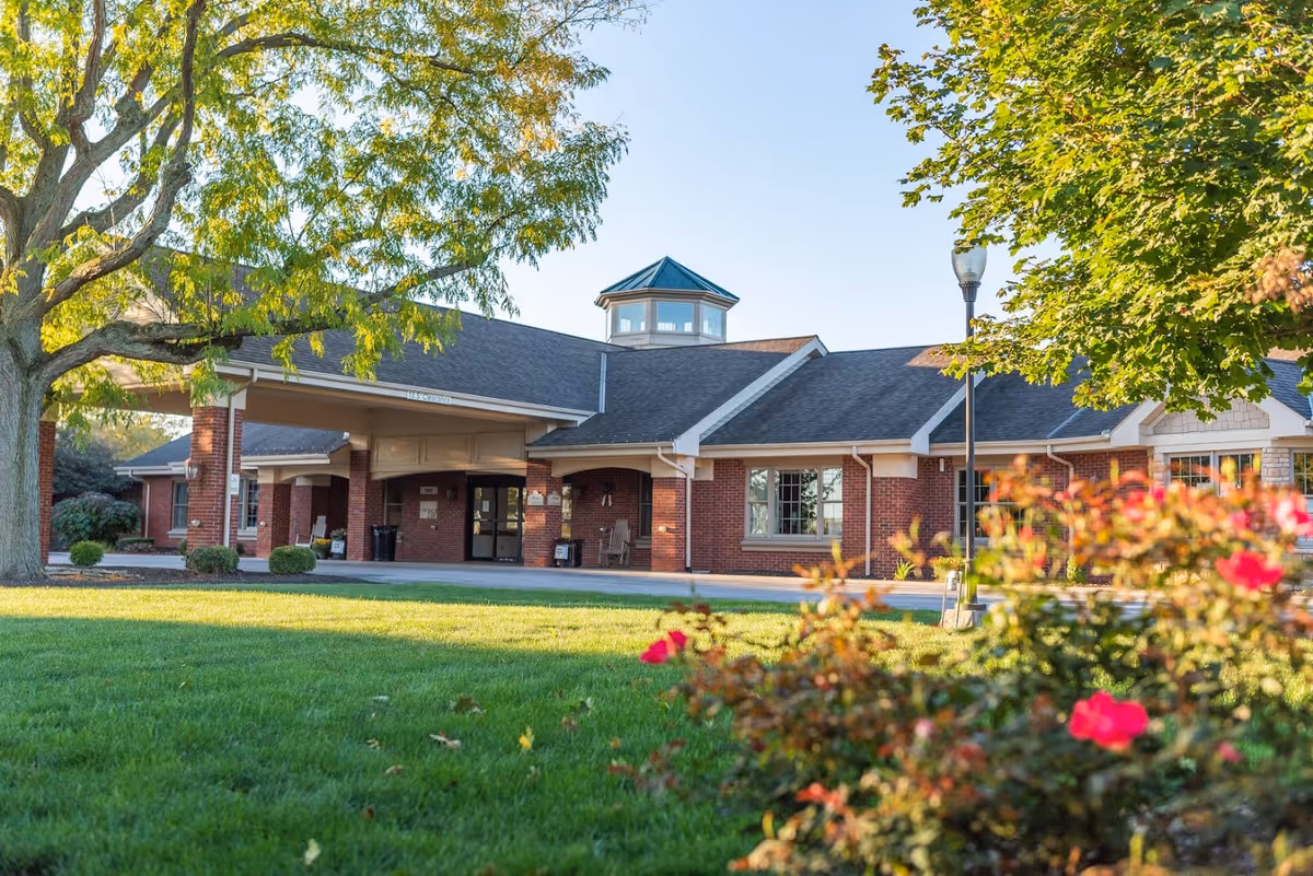Exterior view of a single-story brick building with a covered entrance, surrounded by green grass, trees, and flowering bushes under a clear blue sky.